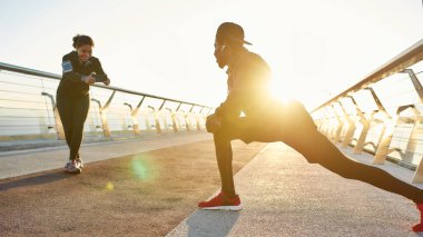 İyi esneme hareketleri. Mutlu Afrikalı kız gülümseyip köprüde gerinen bir adamın fotoğrafını çekiyor. Isınıyor ve kameraya gülümsüyor. Sabah birlikte koşan bir çift.