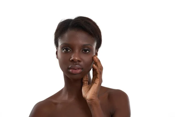 Clean skin. Portrait of a young sensual african woman with naked shoulders touching her face and looking at camera while standing against white background. — Stock Photo, Image