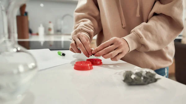 För din avkoppling. Beskuren bild av kvinnan som förbereder cannabisknoppar för slipning med röd marijuanakvarn när hon står i köket. Glasvattenrör eller bong på bordet — Stockfoto