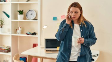 Günlük giysiler içinde genç bir kadın müşteriyle telefonda konuşuyor ve evde çalışırken kahve ya da çay içiyor.