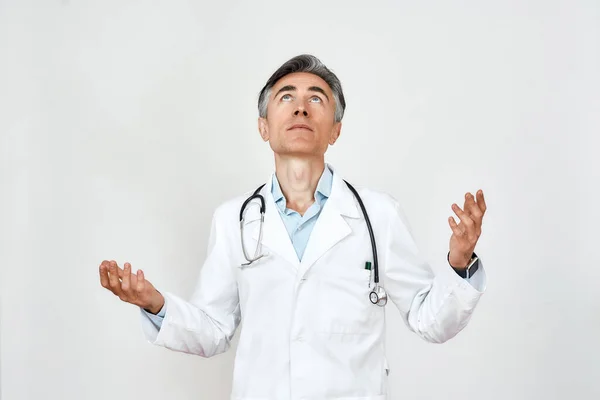 Portrait of upset mature doctor in medical uniform with stethoscope around neck looking up with hope, praying while standing against grey background — Stock Photo, Image