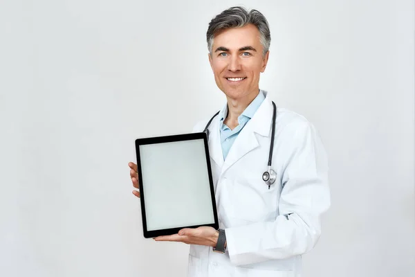 Ευτυχισμένος ώριμος γιατρός με ιατρική στολή με στηθοσκόπιο γύρω από το λαιμό που δείχνει κενό ψηφιακό δισκίο στην κάμερα ενώ στέκεται πάνω σε γκρι φόντο — Φωτογραφία Αρχείου