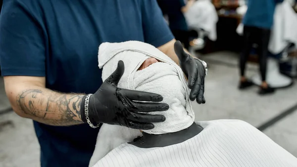 Profesyonel dövmeli berber müşterisinin yüzünü sıcak havluyla kaplıyor, tıraş olmadan önce yüzünü buharla buharlaştırıyor. Berber dükkanındaki adam. — Stok fotoğraf