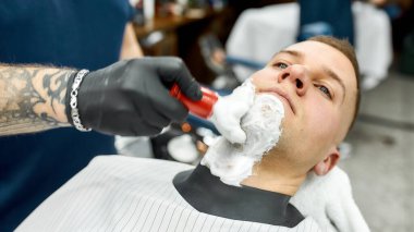 Sakalını kesiyorum. Berber dükkanında çalışırken saç fırçasıyla tıraş köpüğü süren kollu bir berberin kesilmiş fotoğrafı.