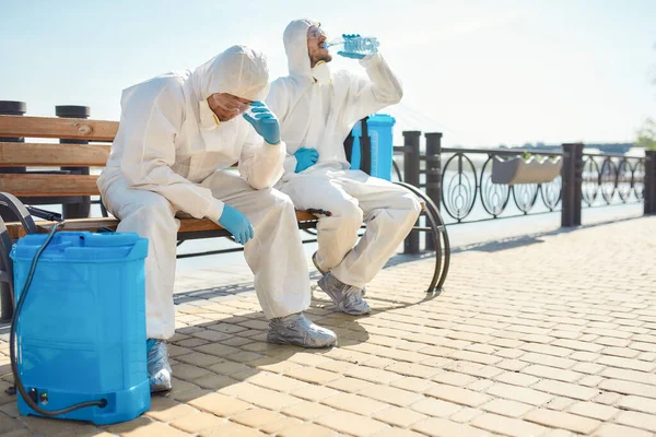 Κουρασμένος. Εξυγιάνσεις, καθαρισμός και απολύμανση της πόλης λόγω της εμφάνισης του ιού Covid19. Άντρες με προστατευτικές στολές και μάσκες κάνουν ένα διάλειμμα κοντά στο ποτάμι. — Φωτογραφία Αρχείου
