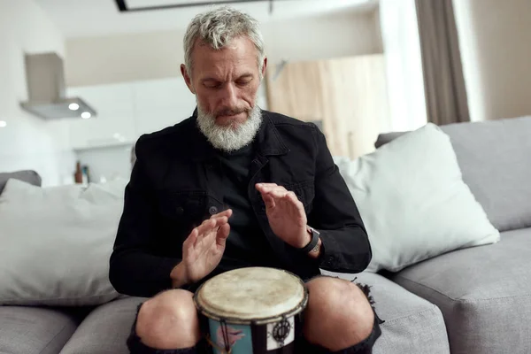 La musica porta armonia. Uomo, artista o compositore barbuto di mezza età che scrive canzoni, suona il rhytm con tamburo djembe, seduto sul divano di casa — Foto Stock