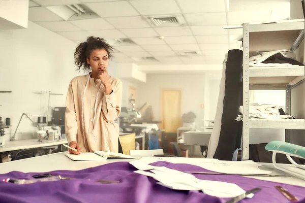 Почувствуйте творческую энергию. Молодая женщина дизайнер одежды опираясь на стол с тканями, текстильными и швейными принадлежностями на нем, при планировании новой коллекции и записи — стоковое фото