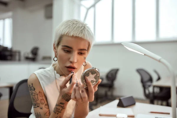 Ajustando su maquillaje. Retrato de una joven y elegante mujer de negocios tatuada rubia que aplica lápiz labial mientras está sentada en el moderno espacio de coworking — Foto de Stock
