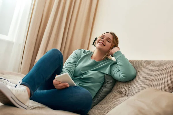 A acalmar. Menina bonita jovem e feliz em roupas casuais usando fones de ouvido, relaxar no sofá e ouvir música — Fotografia de Stock