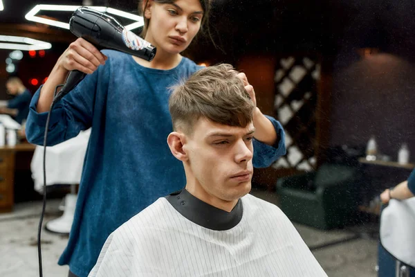 Yeni saç kesimi neredeyse bitti. Modern berber dükkanında ya da kuaförde genç şık bir adam saçını saç kurutma makinesiyle kurutan profesyonel berber kız. — Stok fotoğraf