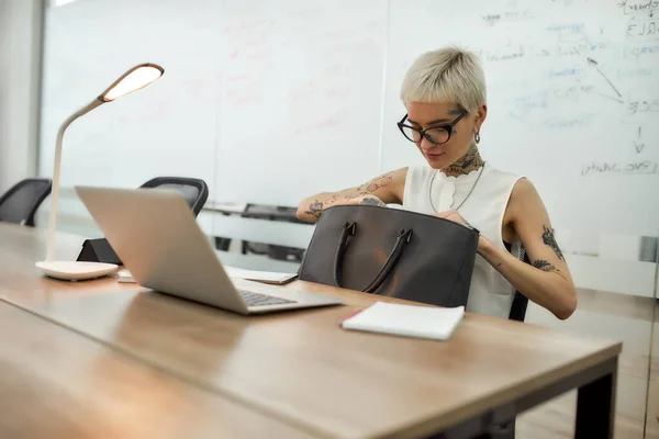 Young attractive blonde tattooed businesswoman with short haircut looking for something in her black handbag while working in the modern office