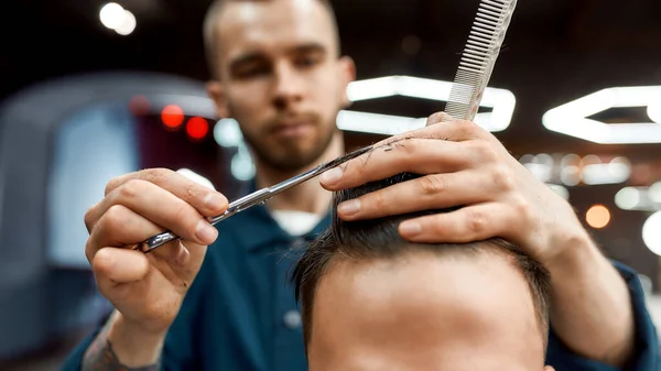Personal stylist. Close up of young professional barber with scissors and hair comb in hands doing haircut for his client in barber shop — Stock Photo, Image