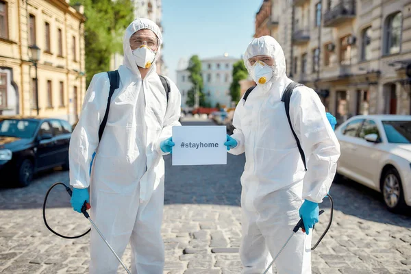 Stay Alert. Cleaning and disinfection of the streets in the city due to the emergence of Covid19 virus. Specialized team in protective suits and masks holding a card with the inscription Stay Home