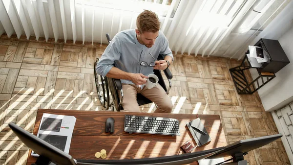 Enjoying fresh coffee. Top view of young businessman or trader in a wheelchair holding his eyeglasses and drinking tea or coffee while sitting at his desk in the bright modern office