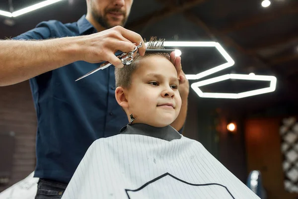 Un piccolo cliente del barbiere. carino ragazzo seduto in poltrona in il barbiere mentre maschio barbiere con forbici in le sue mani fare taglio di capelli per lui — Foto Stock