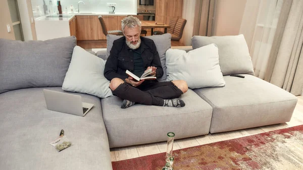 Confort. Homme barbu d'âge moyen lisant un livre assis sur le canapé. Bong ou tuyau d'eau en verre, bourgeons dans un sac en plastique et plus léger près de lui. Concept de légalisation du cannabis et des mauvaises herbes — Photo