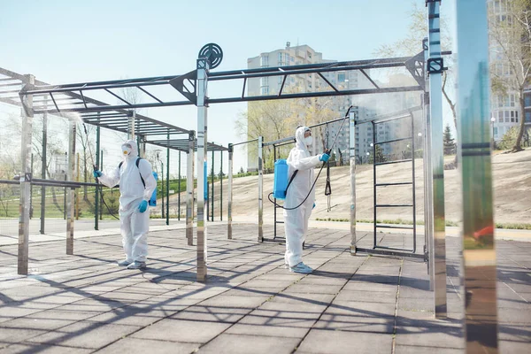 La seguridad es nuestra prioridad. Saneamiento, limpieza y desinfección de la ciudad debido a la aparición del virus Covid19. Equipo especializado en trajes de protección y máscaras en el trabajo cerca del campo de deportes —  Fotos de Stock