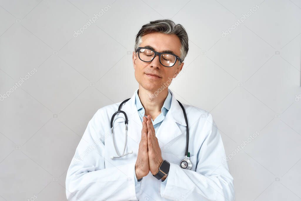 Senior male doctor wearing white coat and glasses, with stethoscope around neck keeping eyes closed and praying for help while standing against grey background