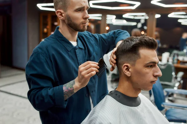 Kolunda tarak tutan ve berber koltuğunda oturan genç yakışıklı adama kuaförlük yapan profesyonel bir berber.