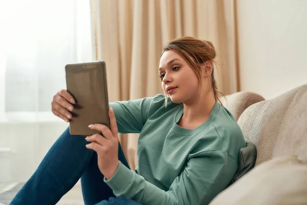 A navegar na Internet. Menina bonita nova em roupas casuais relaxante no sofá na sala de estar em casa e usando tablet digital — Fotografia de Stock