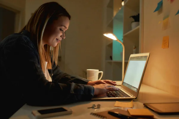 Working in the evening from home. Side view of young smiling woman using laptop, typing on keyboard. Sitting at her home office, working remotely — Stock Photo, Image