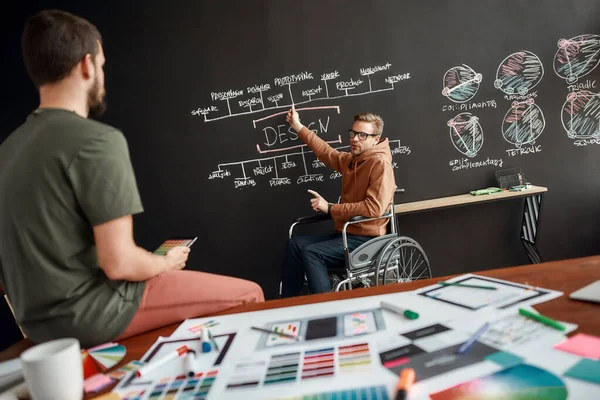 Equipa criativa. Profissional masculino UX UI designer em uma cadeira de rodas explicando algo para seu colega enquanto sentado perto de quadro-negro com apresentação no escritório moderno — Fotografia de Stock