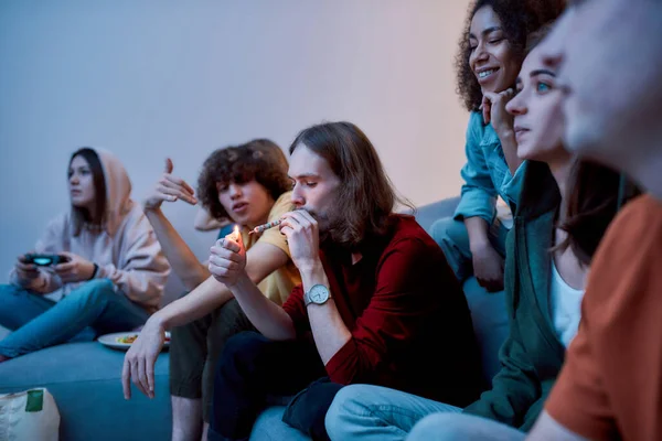 Fuma y relájate. Adolescente joven, hombre encendiendo el cigarrillo de cannabis mientras está sentado con amigos emocionados en el sofá en casa. Jóvenes fumando marihuana, divirtiéndose — Foto de Stock