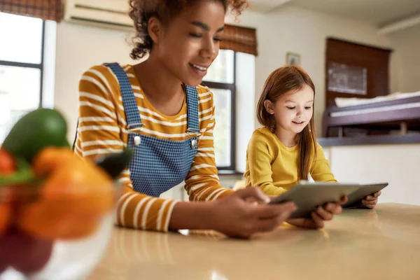 Manera moderna para el cuidado del bebé. Mujer afroamericana niñera y niña linda caucásica leyendo, usando tableta pc, sentada en casa. Educación infantil, actividades de ocio, concepto de cuidado de niños —  Fotos de Stock