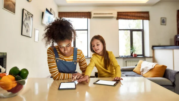 Mujer afroamericana niñera y niña linda caucásica leyendo, usando tableta pc, sentada en casa. Un chico llegando al panel táctil de Nannys. Educación infantil, actividades de ocio, concepto de cuidado de niños —  Fotos de Stock