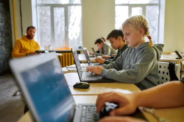 Desarrollar y aprender a través de la diversión y la creatividad. Retrato de colegiala caucásica mirando la pantalla del portátil junto con otros alumnos durante una lección en la escuela inteligente moderna. Enfoque selectivo —  Fotos de Stock