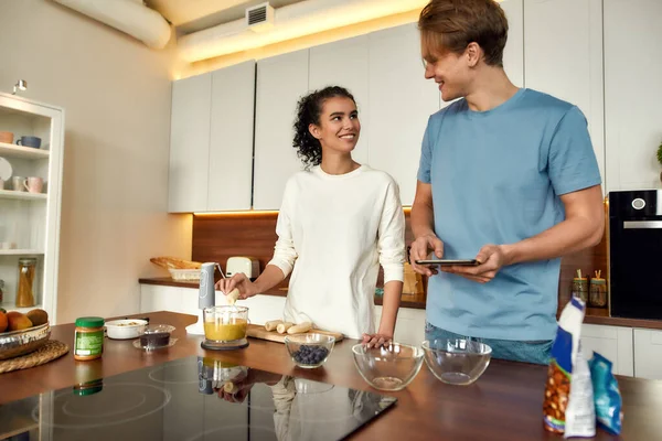 Pareja feliz preparando una bebida saludable, batido juntos en la cocina. Mujer joven poniendo frutas en la licuadora, hombre comprobar la receta con la tableta de PC — Foto de Stock