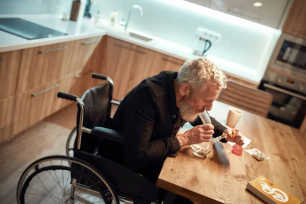 Ik zoek hulp. Gehandicapte man in een rolstoel die cannabis aansteekt in een kom glazen waterpijp of waterpijp in de keuken. Marihuana gereedschap op tafel. Legalisatie, afhankelijkheid van licht drugsconcept — Stockfoto