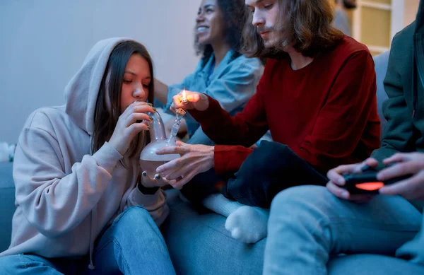 Fumar marihuana y jugar videojuegos. Joven chico bong iluminación bong con marihuana para su amiga, sentarse y relajarse con amigos en el sofá en casa —  Fotos de Stock