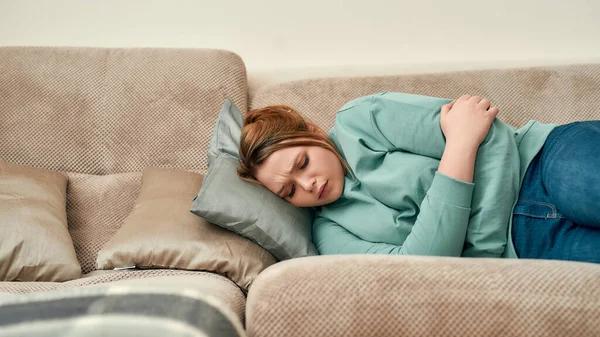 女性健康。年轻而悲伤的女人躺在家里的沙发上，患有经期或腹痛 — 图库照片