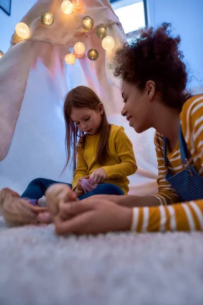 Someone you can trust. African american woman baby sitter entertaining caucasian cute little girl. They are playing with dolls together sitting in wigwam, tent — Stock Photo, Image