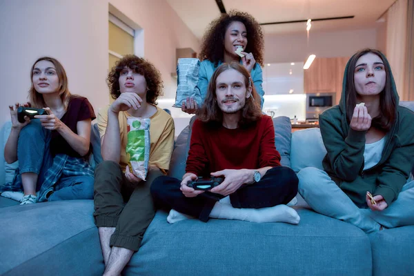 Home entertainment. Young multicultural people playing video games, eating snacks and relaxing together on the couch at home