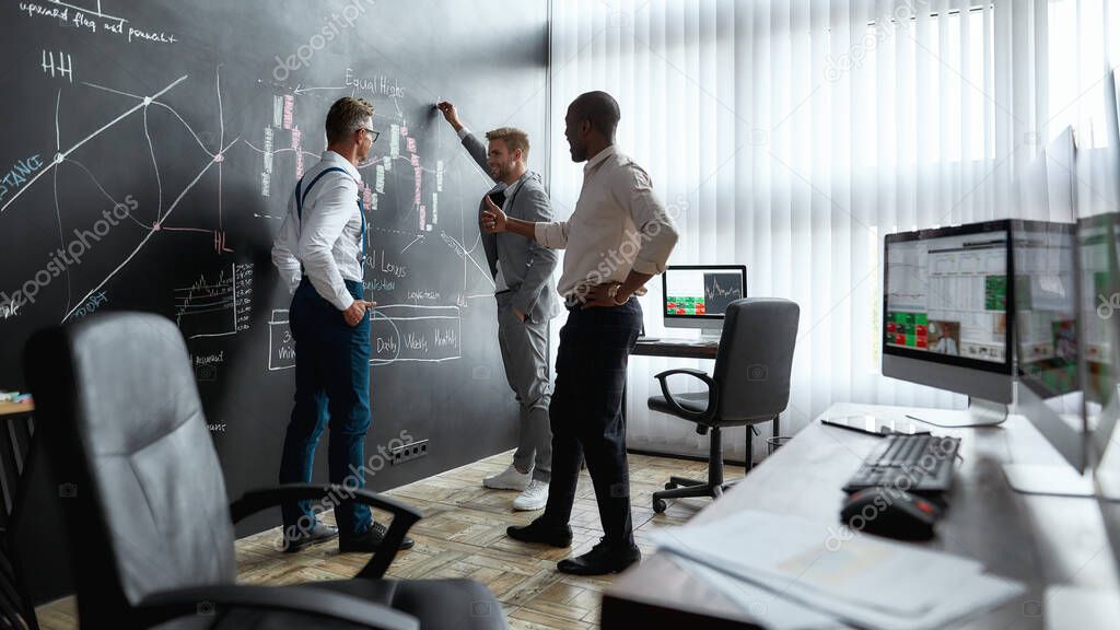 Having advanced level teachers. Full-length shot of diverse employees, traders discussing the strategy of work, while standing near blackboard full of charts. Horizontal shot