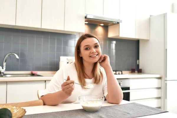 Καλύτερη ζωή με το πρωινό. Νεαρή γυναίκα χαμογελά στην κάμερα ενώ τρώει πλιγούρι βρώμης στην κουζίνα — Φωτογραφία Αρχείου