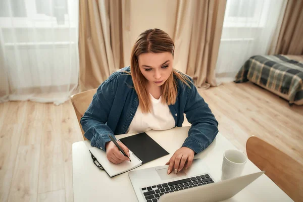 Wolny strzelec. Młoda, kaukaska kobieta w luźnych ciuchach, używająca laptopa i robiąca notatki siedząc w domu, pracuje zdalnie — Zdjęcie stockowe