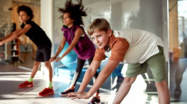 Vücudunu çalıştır. Isınırken kameraya bakan bir çocuğun portresi, spor salonunda diğer çocuklarla birlikte egzersiz yapıyor. Spor, sağlıklı yaşam tarzı, aktif çocukluk konsepti
