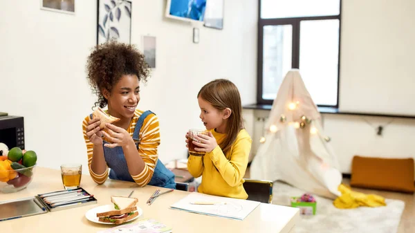 Nutrir a felicidade. afro-americana baby sitter divertido caucasiano bonito menina. Miúdo a beber chá enquanto almoça com a ama. Atividades de lazer, conceito de babá — Fotografia de Stock