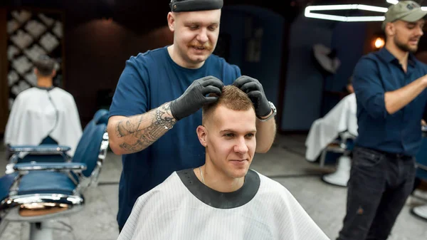 Tolle Arbeit. Stilvoller Friseur mit Tätowierung auf der Hand, der Haarstyling für einen jungen gutaussehenden Kerl macht, der im Stuhl des Friseursalons sitzt. Friseurladen-Konzept. Schuldenschnitt — Stockfoto