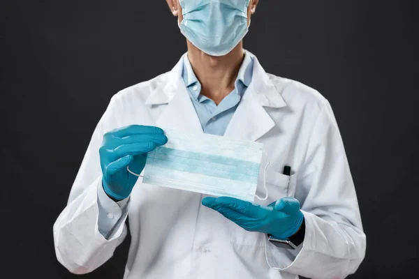 검은 색 배경에 서 있는 동안, 의료 복을 입고 푸른 살균 장갑을 끼고 카메라 앞에서 의료용 마스크를 하고 있는 한 남자 의사의 사진 이 발견되었다 — 스톡 사진