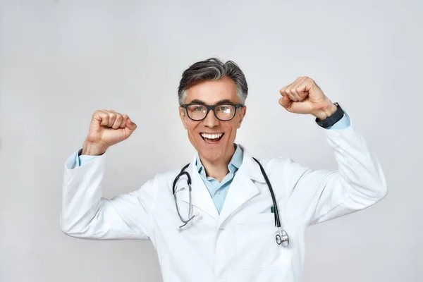 Excited senior caucasian doctor in medical uniform and glasses with arms raised celebrating success, looking at camera and smiling, standing against grey background — Stock Photo, Image