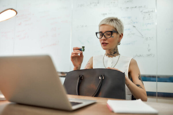 Going to smoke marijuana. Young stylish tattooed businesswoman in eyeglasses holding metal smoking pipe while sitting at her working place in modern office