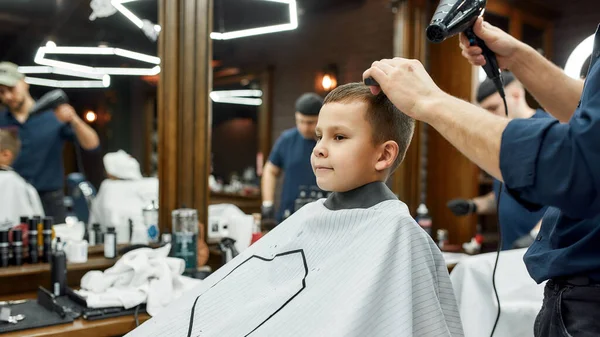 Acconciatura per bambini. Piccolo ragazzo carino seduto in poltrona al barbiere mentre il barbiere asciuga i capelli. Ottenere taglio di capelli — Foto Stock