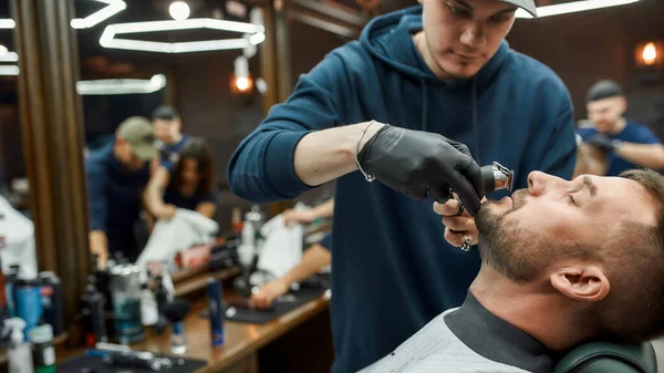 Baffi e toelettatura della barba. Barbiere in guanti neri che taglia i baffi del suo cliente con rasoio elettrico. Un giovanotto in visita dal barbiere. Vista laterale — Foto Stock