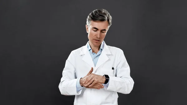 Vigyázz magadra! Érett férfi orvos orvosi egyenruhában mutatja meg, hogyan kell kezet mosni, miközben áll szemben a fekete háttér — Stock Fotó