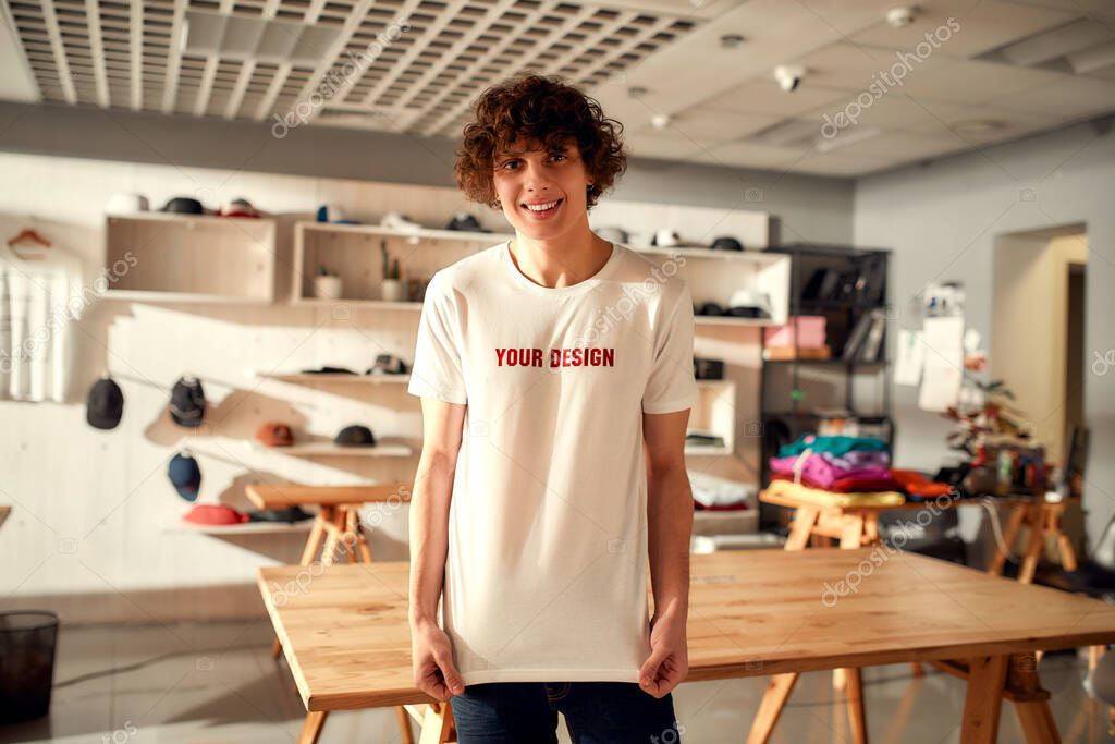 Young man smiling at camera, while trying on printed t shirt, standing in the store. Clothes at custom T-shirt, clothing printing company. Horizontal shot. Selective focus