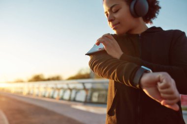 Akıllı telefondan müzik dinleyen kadın koşucuyu kapatın, kolçakla dokunmatik ekran kullanın, sabah güneş doğarken köprüde koşun.